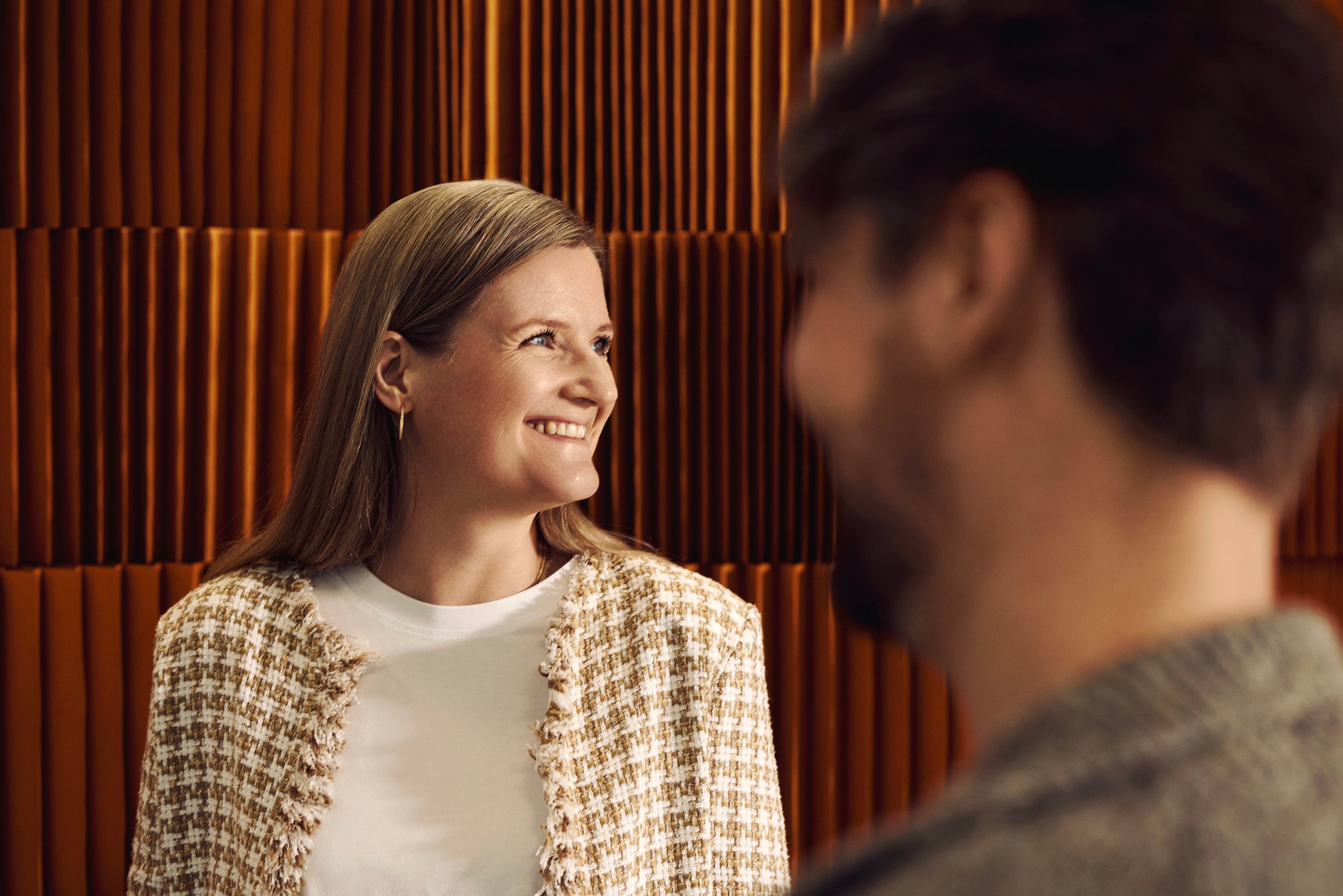 Two people in conversation in front of an orange ribbed wall panel. One smiles and looks to the side.