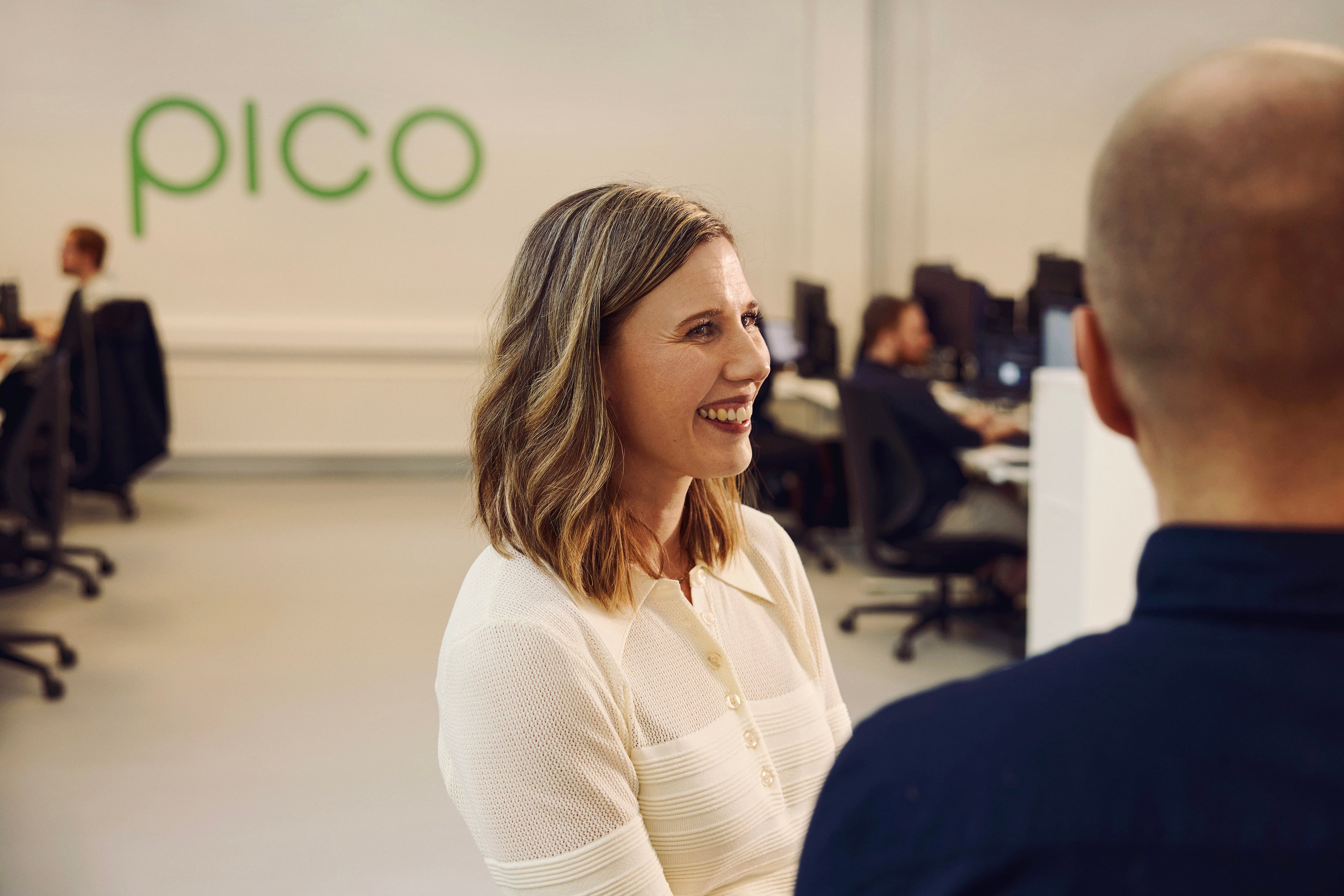 Woman in white shirt talking with colleague at Pico office with employees in background