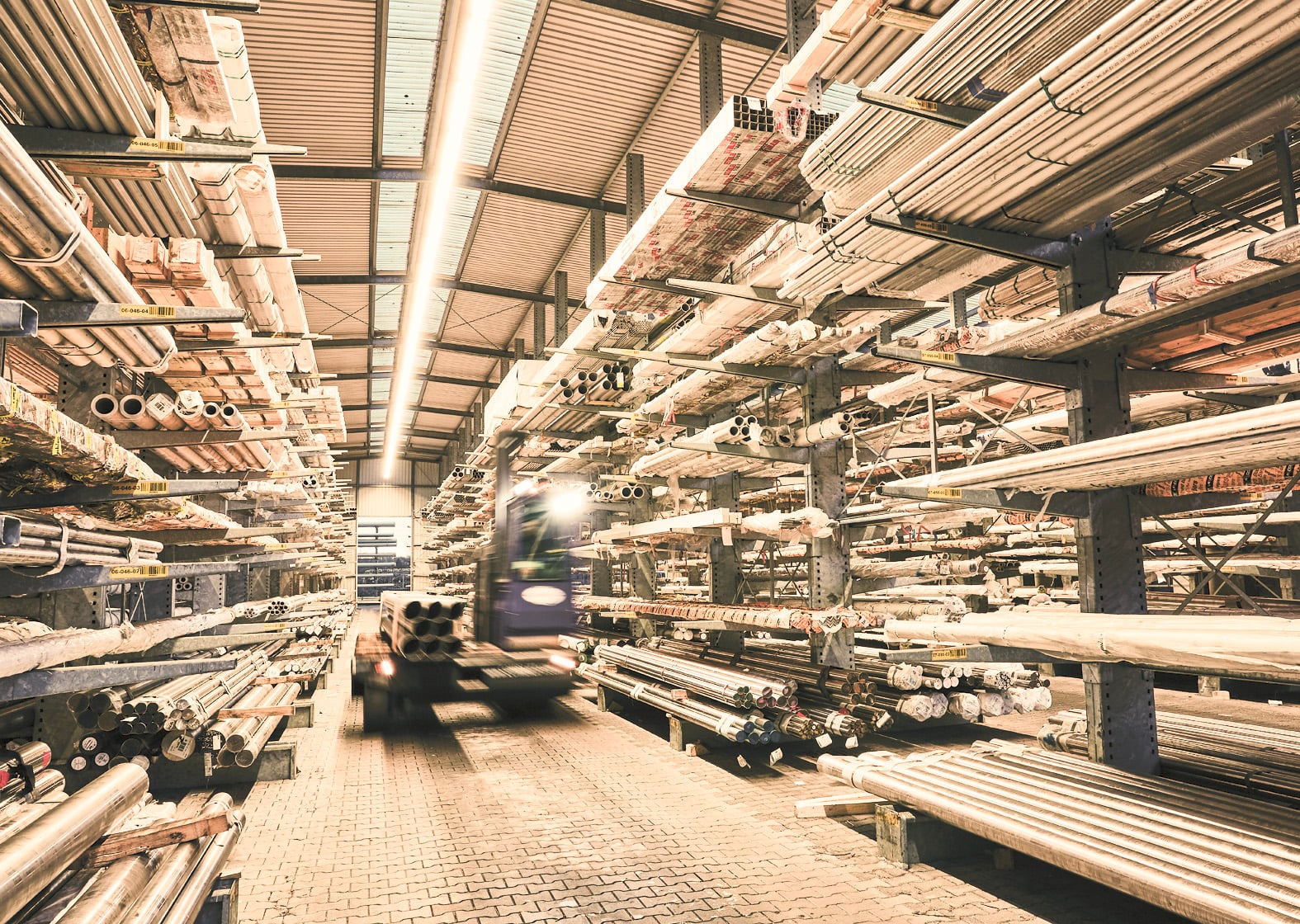 Steel warehouse with pipes and profiles stacked on shelves under industrial roof with forklift in motion.