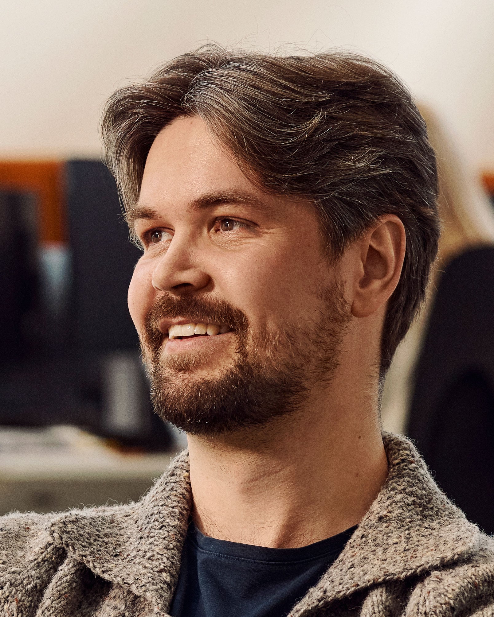 Man with dark hair and beard wearing gray cardigan, smiling to the side