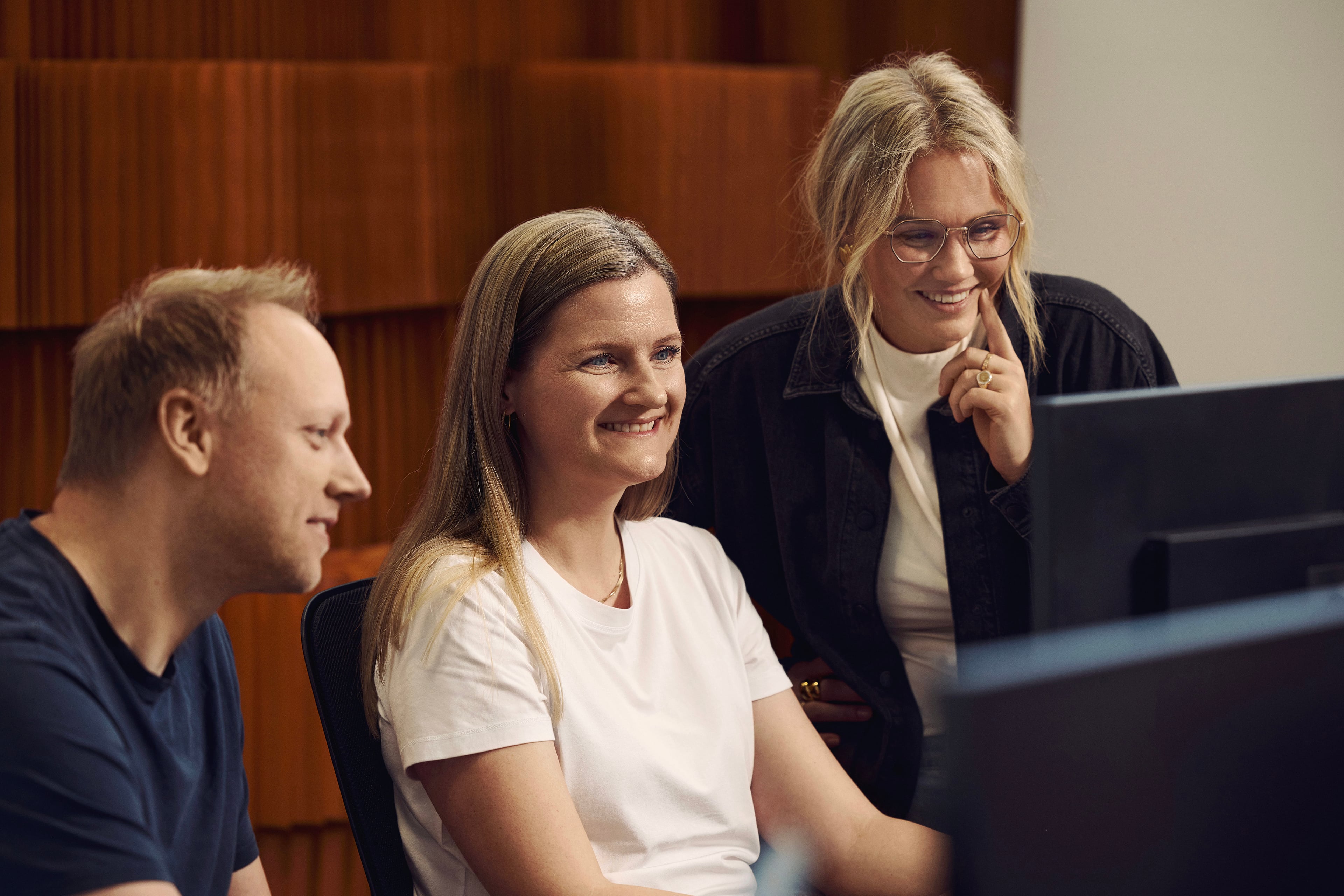 Three people looking at a computer screen in an office with wooden wall paneling in the background